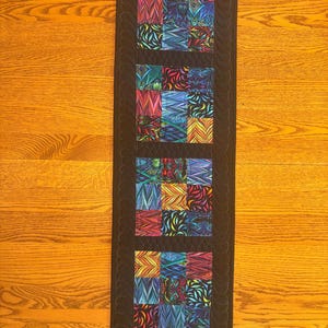 May include: A rectangular quilted table runner featuring a central column of four square blocks, each composed of nine smaller squares with vibrant, multi-colored abstract patterns. The runner is bordered by a solid black fabric with quilted feather motifs.