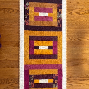 May include: A rectangular quilted table runner with a repeating pattern of rectangles in shades of gold, purple, and white. The design includes floral accents and a white border. The runner is approximately 12 inches wide and 36 inches long.