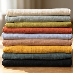Puede incluir: Una pila de nueve piezas de tela dobladas en varios colores sólidos, incluyendo crema, verde salvia, oliva, terracota, azul claro, bronceado, amarillo mostaza, verde azulado oscuro y negro. Cada pieza presenta un patrón tejido texturizado.