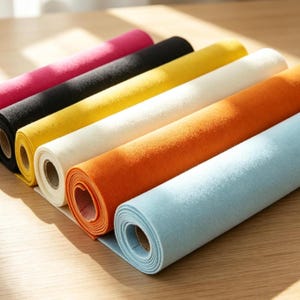 Könnte beinhalten: Eine Sammlung von sechs Stoffrollen in leuchtenden Farben, darunter Magenta, Schwarz, Gelb, Creme, Orange und Hellblau. Jede Rolle hat eine weiche, filzartige Textur und ist um einen stabilen Pappkern gewickelt, die parallel angeordnet sind.