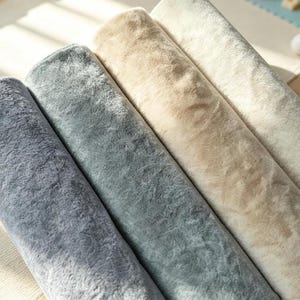 Puede incluir: Cuatro rollos de tela de felpa dispuestos uno al lado del otro. Los rollos presentan una textura suave y difusa en un degradado de colores, incluyendo gris oscuro, gris claro, beige y crema. Cada rollo es cilíndrico y denso.