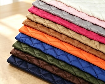 Puede incluir: Una pila de láminas de tela acolchada en varios colores, incluyendo blanco, rosa claro, gris, magenta, beige, naranja, azul, verde oliva, marrón y azul marino. Cada lámina presenta un patrón de costura en forma de diamante uniforme.