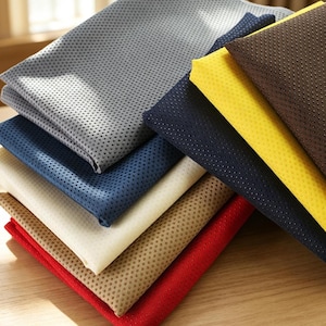 Puede incluir: Una pila de láminas de tela dobladas en varios colores, incluyendo gris, azul marino, crema, beige, rojo, amarillo y marrón. Cada pieza de tela presenta una superficie texturizada uniforme cubierta con pequeños puntos en relieve espaciados.