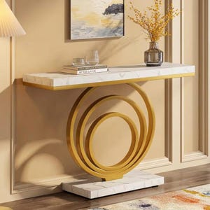 Può includere: Un tavolo console con piano e base rettangolari in marmo bianco. La struttura di supporto è composta da tre anelli metallici dorati concentrici. Sul tavolo sono presenti un vaso di vetro con fiori gialli e una pila di due libri.