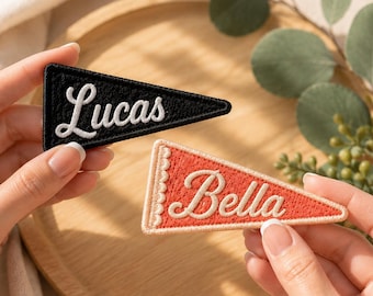May include: Two triangular pennant-shaped embroidered patches. One patch is black with white stitched borders and the name Lucas in white script. The second patch is orange with cream stitched borders and the name Bella in cream script.