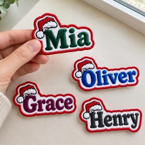 Puede incluir: Cuatro parches bordados con nombres que presentan un icono de gorro de Papá Noel rojo y blanco sobre los nombres Mia en verde, Oliver en azul, Grace en morado y Henry en negro. Cada parche tiene un borde blanco y un contorno rojo.