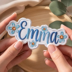 Può includere: Una toppa ricamata bianca con bordo smerlato che riporta il nome Emma in corsivo blu. La toppa è decorata con quattro piccoli fiori azzurri con centri arancioni, disposti attorno alla scritta su uno sfondo in tessuto bianco.