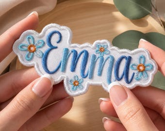 May include: A white embroidered patch with a scalloped edge featuring the name Emma in blue cursive script. The patch is decorated with four small light blue flowers with orange centres, arranged around the lettering on a white fabric background.