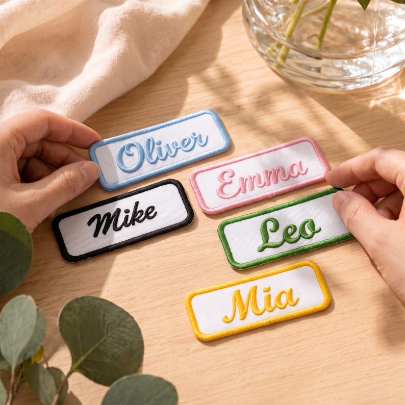 May include: Five rectangular embroidered name patches with white fabric centres and coloured borders. The patches feature the names Oliver in blue, Emma in pink, Mike in black, Leo in green, and Mia in yellow, all in a cursive script font.