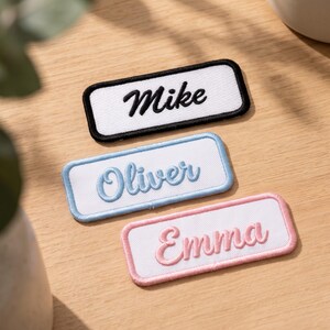 May include: Three rectangular embroidered name patches with white fabric centres. The top patch has a black border and the name Mike in black script. The middle patch has a light blue border and the name Oliver in light blue script. The bottom patch has a pink border and the name Emma in pink script.