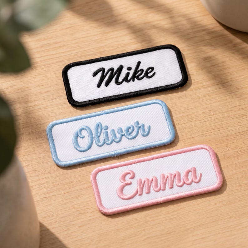 May include: Three rectangular embroidered name patches with white fabric centres. The top patch has a black border and the name Mike in black script. The middle patch has a light blue border and the name Oliver in light blue script. The bottom patch has a pink border and the name Emma in pink script.