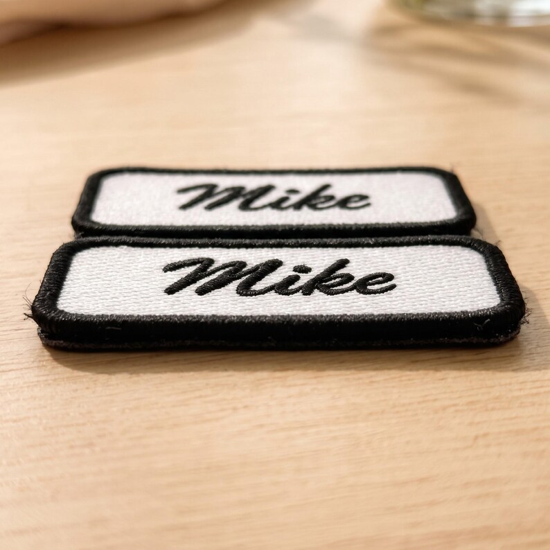 May include: Two rectangular embroidered patches with a white background and a thick black border. Each patch features the name Mike stitched in black cursive script. The patches are stacked one in front of the other on a light wooden surface.