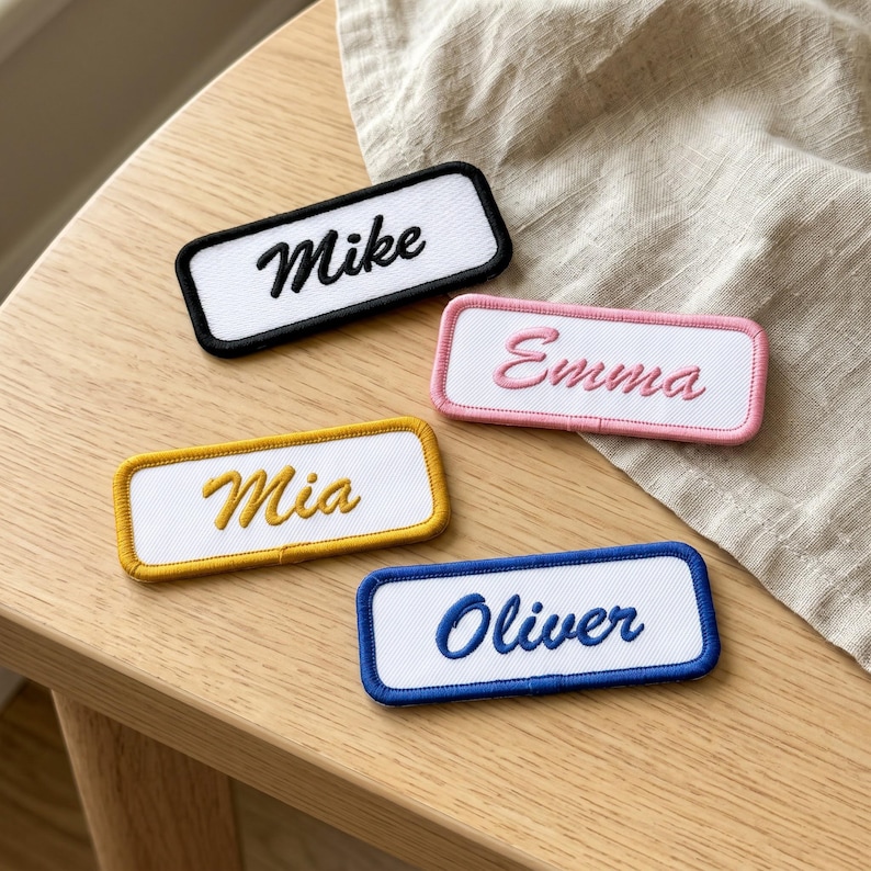 May include: Four rectangular embroidered name patches with white fabric centres and coloured borders. The patches feature the names Mike in black, Emma in pink, Mia in yellow, and Oliver in blue, all written in a cursive script font.