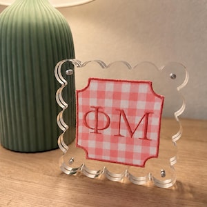 May include: A clear acrylic square frame with scalloped edges featuring a pink and white gingham fabric insert. The fabric is embroidered with the Greek letters Phi and Mu in a matching pink thread. The frame is held together by four metal screws.