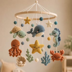 Può includere: Una giostrina sospesa con creature marine all'uncinetto, tra cui una balena, una tartaruga, un polpo, un cavalluccio marino, un granchio e una conchiglia con perla. Le figure sono appese a un telaio circolare in legno con fili bianchi.