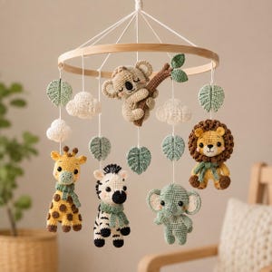 May include: A hanging nursery mobile featuring a circular wooden frame with crochet figures of a koala on a branch, a giraffe, a zebra, a lion, and an elephant. The mobile includes white crochet clouds and sage green crochet leaves suspended by white strings.