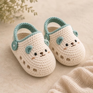 May include: A pair of cream-colored crochet baby shoes featuring a bear face design with small teal ears, embroidered black eyes and nose, and a teal heel strap secured by a matching teal button on each side of the footwear.