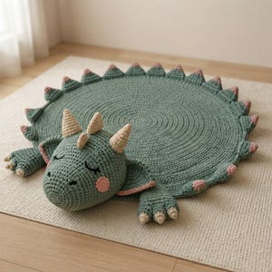 May include: A round, sage green crocheted rug shaped like a sleeping dinosaur. The rug features a textured circular body with a row of small, pink-tipped triangular spikes along the edge, a stuffed dinosaur head with beige horns, and two front paws.