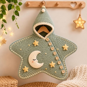 May include: A sage green crocheted star-shaped hooded blanket featuring a cream-colored crescent moon with a sleeping face, several small yellow crocheted stars, and a row of eight light brown wooden buttons along the diagonal front opening.