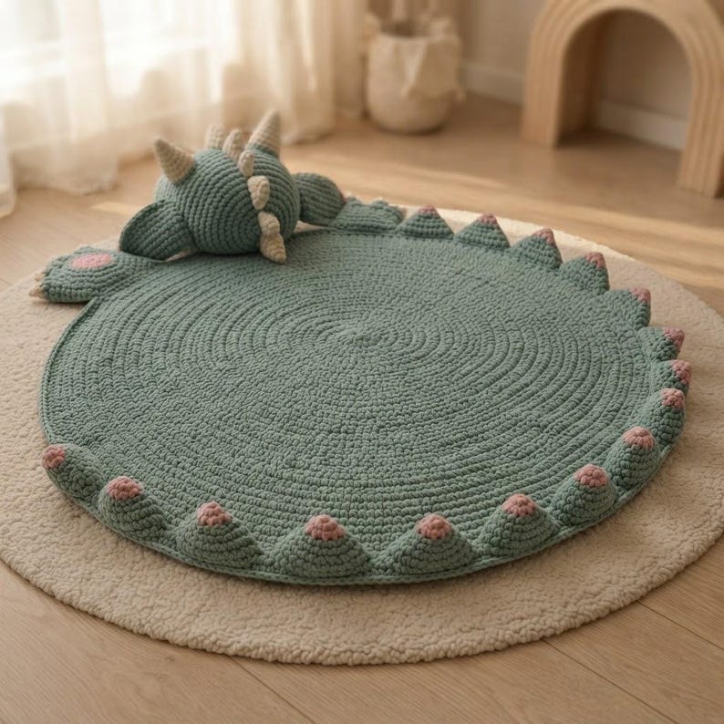 May include: A circular, sage green crocheted rug featuring a three-dimensional dinosaur head, arms, and tail. The perimeter is adorned with a series of small, sage green triangular spikes, each tipped with a soft pink accent, creating a textured border.