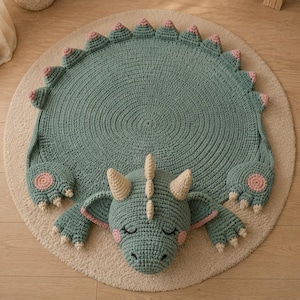 May include: A round, sage green crocheted rug shaped like a dragon. The rug features a textured body with a row of triangular spikes along the edge, two front paws with pink circular pads, and a central dragon head with cream-coloured horns.
