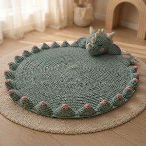 May include: A round, sage green crocheted rug featuring a three-dimensional dragon head with closed eyes, small horns, and pink cheeks. The perimeter of the rug is decorated with a series of small, pointed, sage green spikes with pink tips.