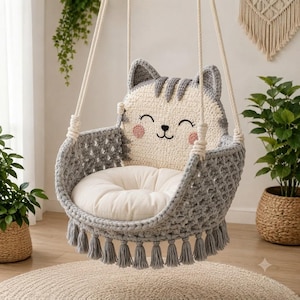 May include: A hanging chair featuring a crocheted cat face backrest in cream and gray with pink cheeks. The seat is a round white cushion nestled within a gray crocheted basket structure with a lattice pattern and gray fringe tassels at the base.