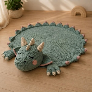 May include: A round, sage green crocheted rug shaped like a sleeping dragon. The rug features a textured, circular body with soft pink spikes along the edge, a plush dragon head with cream-coloured horns, closed eyes, and pink cheeks.