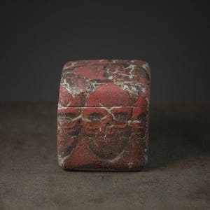 Puede incluir: Una caja de forma cuadrada con un patrón repetitivo de calaveras en tonos rojo, marrón y blanco. La superficie tiene una apariencia texturizada y agrietada con motivos de calaveras visibles en la tapa y los lados del recipiente.