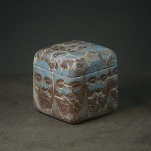 Puede incluir: Una caja cuadrada de cerámica con tapa, que presenta una superficie texturizada con un patrón agrietado en tonos azul claro y marrón tierra. La caja tiene esquinas redondeadas y un acabado mate, mostrando un diseño abstracto repetido.