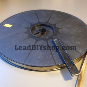 Peut inclure: Une bobine en plastique noir dotée d'un moyeu central et de rayons radiaux, avec un petit autocollant rectangulaire jaune sur la surface supérieure. Un clip en plastique noir est fixé au bord et une bande métallique dépasse de la bobine.