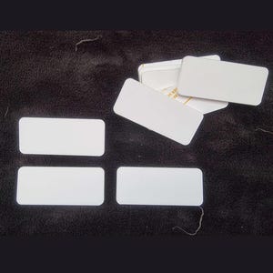 May include: A collection of rectangular white labels with rounded corners. The labels are arranged in a group, with some lying flat and others stacked in a small pile. The surface of each label is plain white, and they feature a clean, simple design.