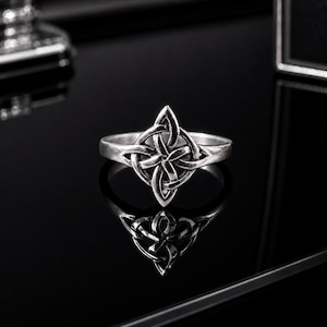 May include: A silver-toned ring featuring a central knotwork design with four pointed petals. The metal has a polished finish with darkened recessed areas to highlight the intricate, interlaced pattern of the symmetrical knot motif.