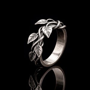 May include: A silver-toned metal ring featuring a band designed to resemble intertwined vines with multiple small, pointed leaves. The metal has an oxidized, darkened finish in the recessed areas, highlighting the texture of the leaves and stems.