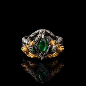 May include: A ring featuring two intertwined dark silver snakes with textured scales, surrounding a central marquise-cut green gemstone. The snakes are positioned above gold-toned leaf-like accents, with small green stones set as eyes on the snake heads.