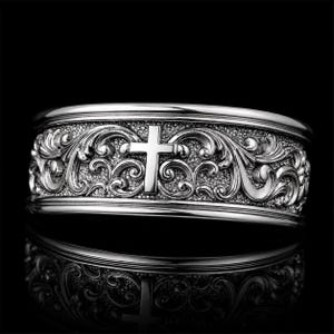 May include: A silver-coloured ring featuring an intricate, raised scrollwork pattern across the entire band. A prominent, centred cross emblem is integrated into the detailed metal relief, which is set against a textured, darkened background.