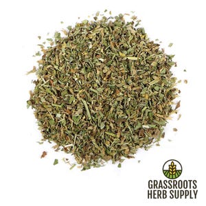 May include: A close-up of a pile of dried, green, and brown herbs. The herbs are loose and appear to be chopped or shredded. The herbs are on a white background.