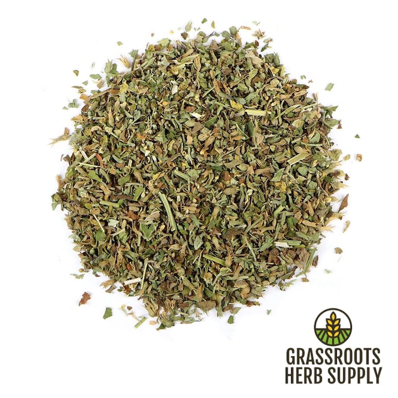 May include: A close-up of a pile of dried, green, and brown herbs. The herbs are loose and appear to be chopped or shredded. The herbs are on a white background.