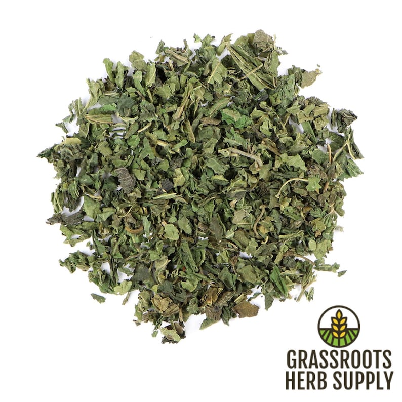May include: Dried nettle leaves, a green herbal ingredient, in a loose pile on a white background. The image is labeled "Grassroots Herb Supply".