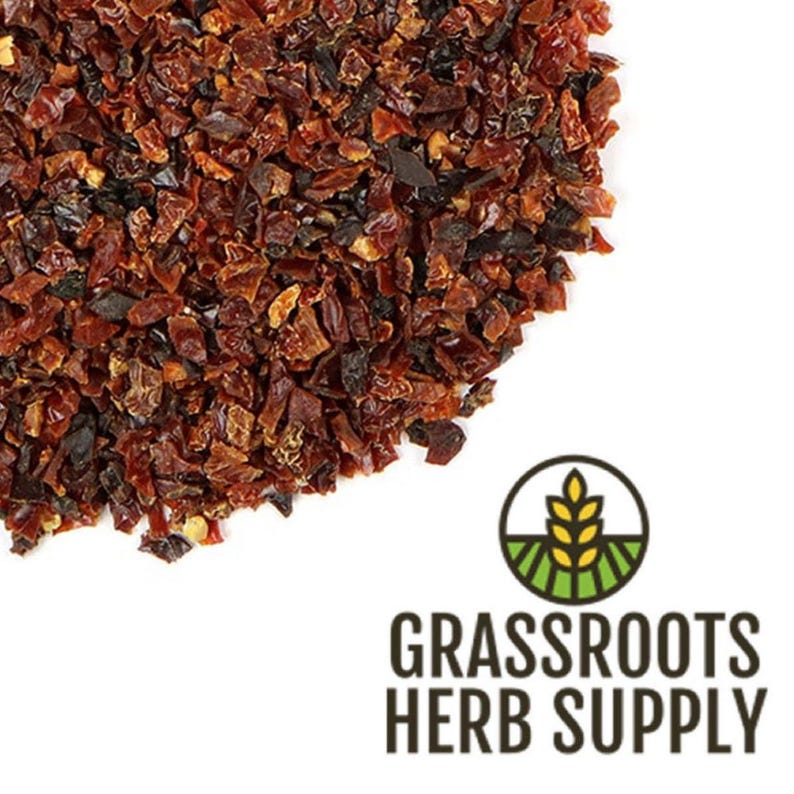 May include: Dried red pepper flakes, a vibrant red color, scattered on a white surface. The flakes are small and irregular in shape, with a slightly glossy texture. The image is taken from a close-up perspective, showcasing the detail of the flakes.