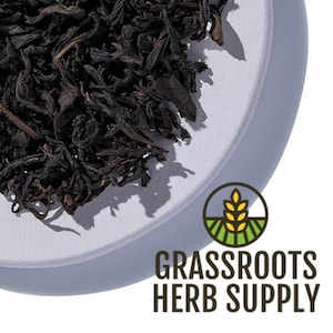 May include: Loose black tea leaves on a white ceramic plate. The tea leaves are dark brown and have a slightly curled shape. The plate is round and has a slightly raised edge. The text "GRASSROOTS HERB SUPPLY" is printed in brown letters on a white background.