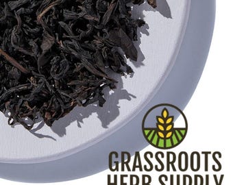 Pode incluir: Folhas de chá preto soltas em um prato de cerâmica branco. As folhas de chá são de cor marrom escura e têm uma forma ligeiramente encurvada. O prato é redondo e tem uma borda ligeiramente elevada. O texto "GRASSROOTS HERB SUPPLY" está impresso em letras marrons sobre fundo branco.