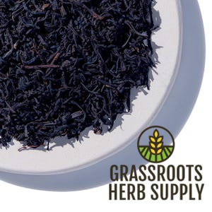 May include: Loose black tea leaves in a white ceramic bowl. The bowl is on a white surface. The text "GRASSROOTS HERB SUPPLY" is visible in the lower right corner of the image.