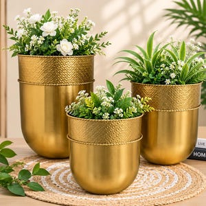 Op de afbeelding: Drie goudkleurige metalen plantenbakken met afgeronde bodems en gestructureerde bovenranden. Elke bak bevat een verscheidenheid aan groen blad en kleine witte bloemen. De plantenbakken zijn in een groep geplaatst, met twee hogere achteraan en een kortere vooraan.