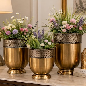Puede incluir: Tres maceteros de metal en tono latón con un patrón festoneado texturizado alrededor del borde superior. Cada maceta contiene una variedad de flores artificiales, incluyendo margaritas blancas, flores rosas y lavanda morada.