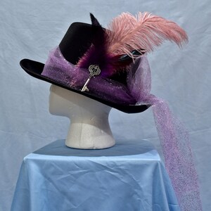 May include: A black top hat with a purple and pink feather and a key charm. The hat is decorated with a purple and pink tulle veil.
