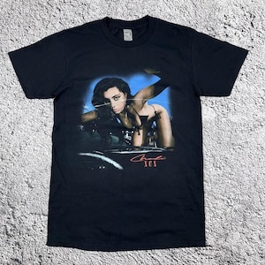 May include: A black short-sleeve t-shirt featuring a graphic print of a person leaning over a car hood against a blue background. The text Charli XCX is printed in red script and block letters below the image on the front of the garment.
