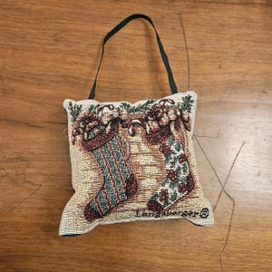 May include: A square tapestry ornament featuring a woven design of two Christmas stockings hanging from a mantel with greenery. The fabric has a beige background with red, green, and white details. A black cord is attached for hanging. Longaberger text.