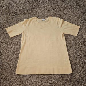 May include: A short-sleeved, light yellow knit sweater featuring a round neckline and a straight hem. The garment is constructed with a fine-gauge knit fabric, showcasing a solid, uniform color throughout the body and sleeves with ribbed trim.