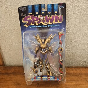 Op de afbeelding: Todd McFarlane's Manga Spawn Ultra-Action-figuur in originele verpakking. Het figuur heeft een goudkleurig en paars pantser, doorschijnende vleugels en accessoires waaronder een staf en een zwaard. De verpakking bevat de tekst Manga Spawn.